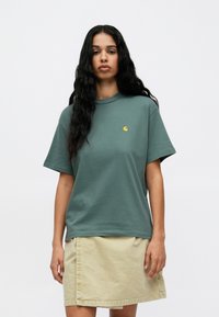 Πράσινο βαμβακερό μπλουζάκι με στρογγυλή λαιμόκοψη και κοντά μανίκια, με μικρό κίτρινο λογότυπο στην αριστερή πλευρά του στήθους. Φορεμένο με μια μπεζ φούστα.