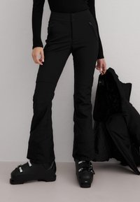 Person iført svarte skibukser og skistøvler, holder en svart vinterjakke med pelsforet innside mot en ensfarget grå vegg.