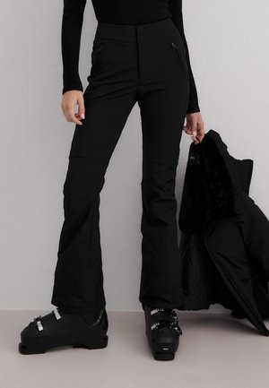 Persoon gekleed in zwarte skibroek en skischoenen, houdt een zwarte winterjas met een bontgevoerde binnenkant vast tegen een effen grijze muur.