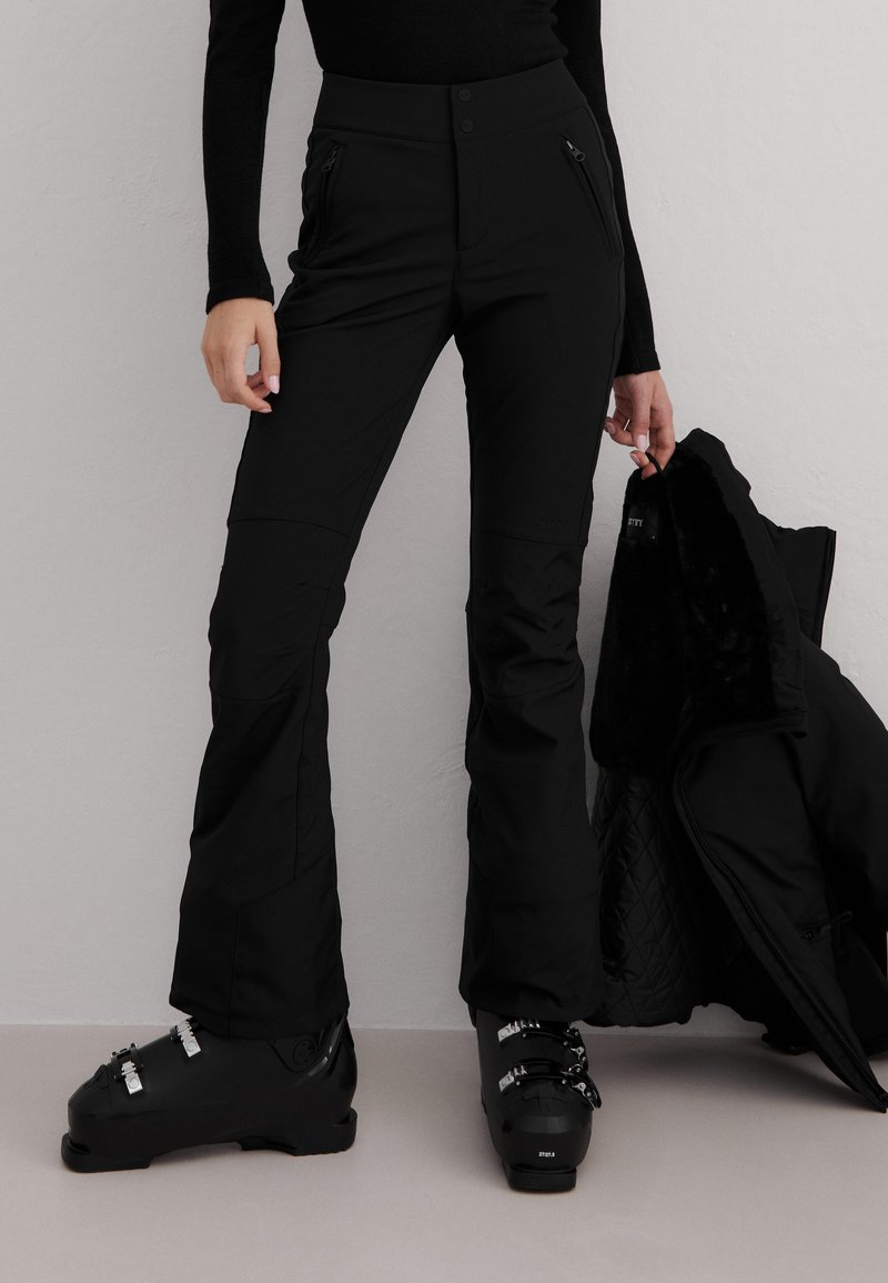 Person iført svarte skibukser og skistøvler, holder en svart vinterjakke med pelsforet innside mot en ensfarget grå vegg.