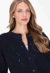 Navy kötött kardigán texturált mintával, kerek nyakkivágással és négy gombos záródással. Laza szabású és puha gyapjú anyagból készült.
