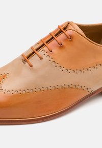 Scarpa in pelle marrone chiaro caratterizzata da una texture liscia, perforazioni rotonde lungo le cuciture e lacci arancioni. Design della punta leggermente affusolato.