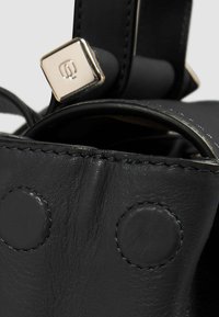 Bolso de cuero negro con una textura suave, herrajes de metal cuadrados y acentos circulares decorativos cosidos en los laterales.