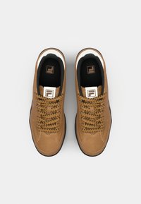 Zapatillas de gamuza marrón con cordones negros, detalles blancos en el collar y una etiqueta de logo en la lengua. Suela de goma plana, diseño clásico de cordones.