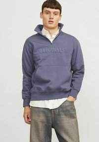 Felpa viola con colletto a metà zip, caratterizzata da una tasca frontale e testo in rilievo "ORIGINALS". Indossata sopra una camicia bianca con colletto.
