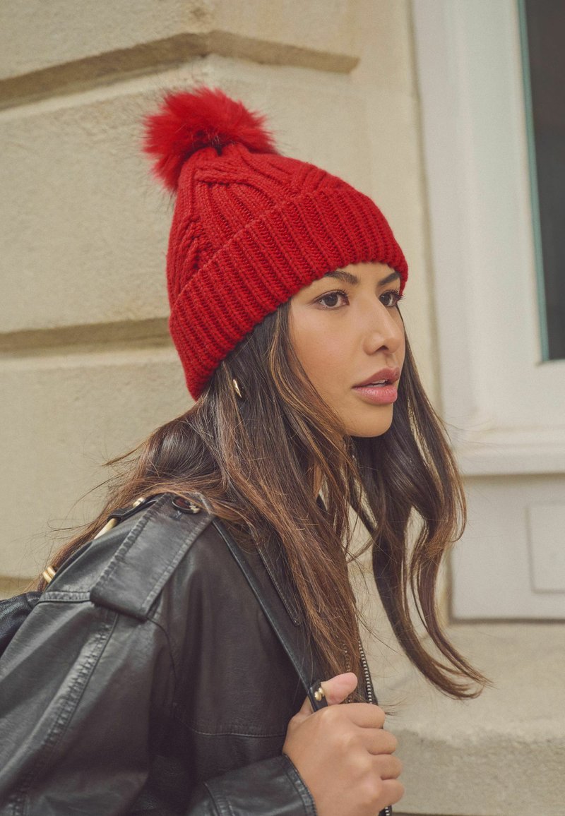 Bonnet tricoté rouge avec un pompon sur le dessus, présentant une texture côtelée et un motif en tresses, porté avec une veste en cuir noire.