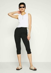Top cropped branco com bainha rendilhada e três botões, combinado com leggings capri pretas e sandálias deslizantes bege. Os acessórios incluem óculos de sol.