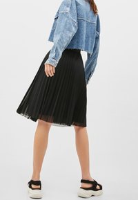 Kvinna i en kort blå jeansjacka, svart plisserad midikjol och svarta sandaler med tjocka vita sulor, stående mot en enhetlig bakgrund.