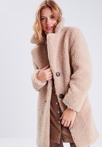 Hellbeige Teddy-Mantel mit hohem Kragen, doppelt besetzten Knöpfen und einem weichen, strukturierten Material. Getragen über einem braunen, knöpfbaren Kleid.