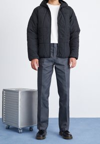Zwarte gewatteerde pufferjack met een capuchon, witte t-shirt eronder, grijze op maat gemaakte broek en zwarte schoenen. Vergezeld door een metalen krat.
