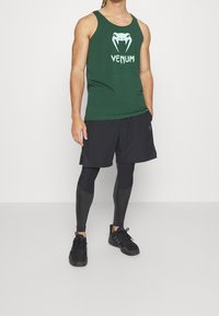 Top senza maniche verde con il logo bianco di Venom, abbinato a pantaloni corti neri e sneaker nere. Struttura testurizzata, vestibilità atletica con proprietà traspiranti.