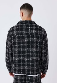 Man met kort haar in een zwart-witte geblokte jas en bijpassende broek, staande met zijn rug naar de camera tegen een effen achtergrond.