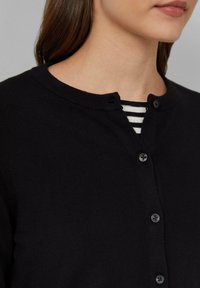 Cardigan noir en tricot avec un col arrondi, présentant un devant à boutons et une texture subtile. Porté par-dessus une chemise à rayures à col ras du cou.