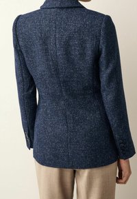 Blazer texturizado azul marino con hombros estructurados, dos botones en cada puño y una silueta entallada. La parte trasera presenta una costura central para dar forma.