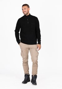 Maglione nero lavorato a maglia con colletto alto, tre bottoni neri e tasca sul petto; abbinato a pantaloni cargo beige e stivali neri.