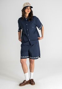 Mørkeblå kortærmet skjorte og matchende shorts med hvid blomsterbroderi, parret med hvide sokker og brune sko, afsluttet med en beige kasket.