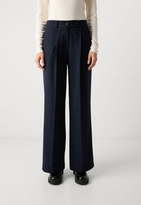 Pantaloni a gamba larga a righe navy con pieghe frontali, due bottoni in vita e una texture liscia. Abbinati a scarpe platform nere.