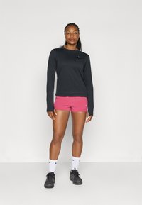 Zwarte langemouwtop met een klein wit logo, gecombineerd met roze shorts. Het model draagt witte sokken en zwarte sportschoenen. Eenvoudig, modern ontwerp.