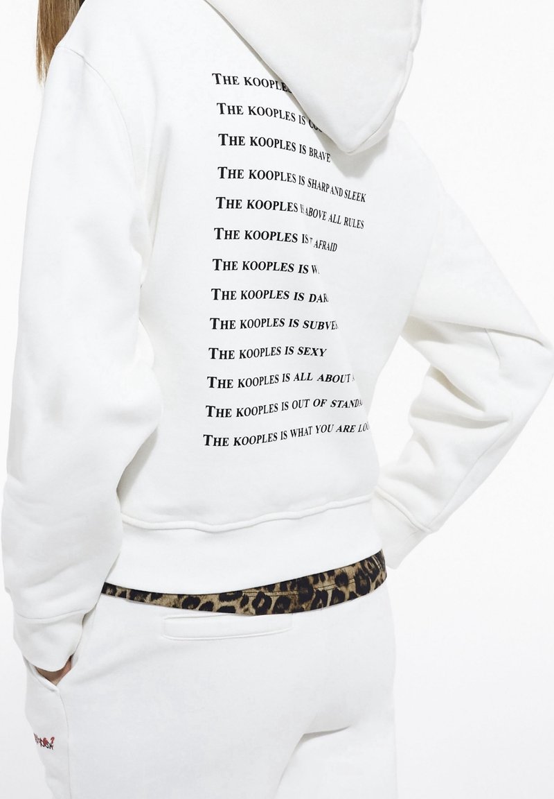 Personne portant un sweat à capuche blanc avec du texte noir énumérant des phrases sur "The Kooples" dans le dos, associé à un pantalon blanc et une ceinture à imprimé léopard.