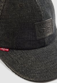 Gorra de pana negra con visera plana, que presenta una superficie texturizada y un parche rectangular de piel con el logotipo de Levi Strauss.