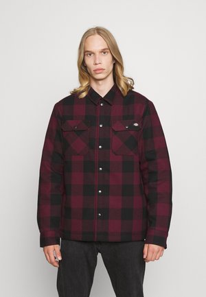 Chaqueta de franela a cuadros rojos y negros con botones, dos bolsillos en el pecho y un corte relajado, hecha de tela texturizada.