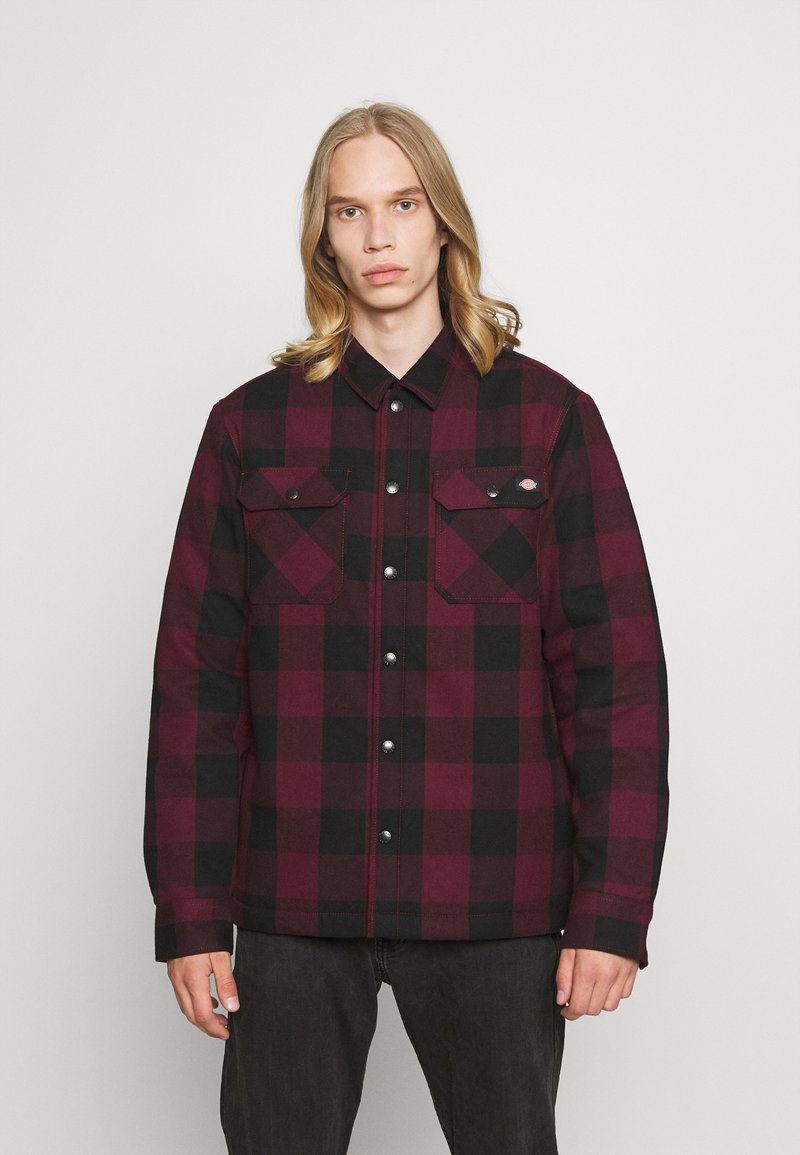 Veste en flanelle à carreaux rouges et noirs avec des boutons, deux poches poitrine et une coupe décontractée, fabriquée à partir d'un tissu texturé.