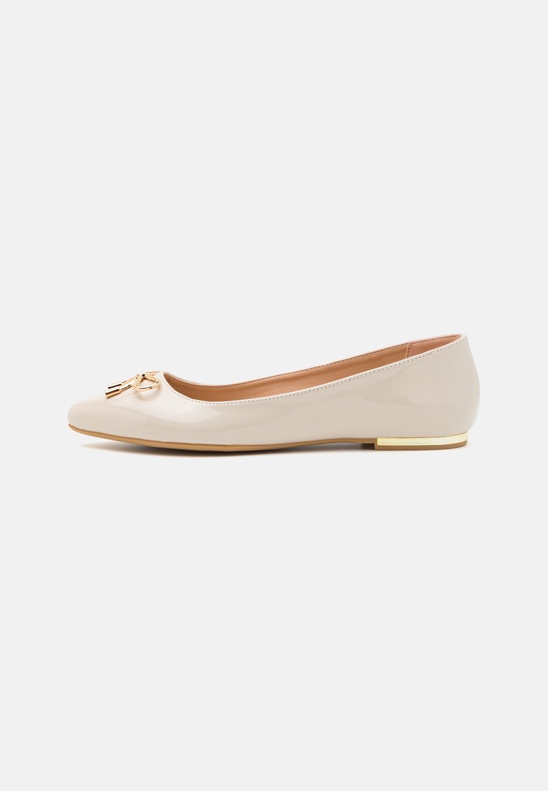 Chaussure plate en cuir verni beige avec un petit talon doré et un pompon doré décoratif sur l’orteil arrondi, vue de côté.