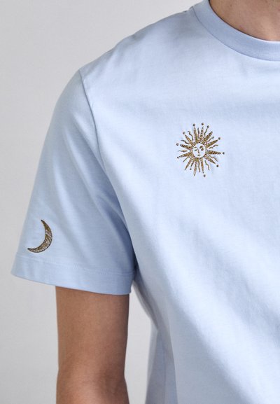 Ljust blå bomulls-t-shirt med guldbroderi av en sol på bröstet och en halvmåne-detalj på ärmen. Kortärmad, rund halsringning.
