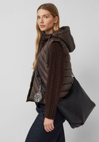 Frau mit langen blonden Haaren, die eine braune gepolsterte Weste und einen Pullover trägt, dabei eine große schwarze Tasche und ein kleines, kreisförmiges Täschchen im Leopardenmuster hält.
