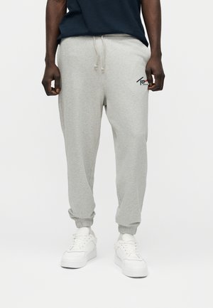 Mand iført lysegrå joggingbukser med elastiske manchetter og hvide sneakers, stående mod en ensfarvet baggrund.