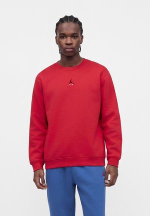 Man with braided hair wearing a red sweatshirt with black logo and blue sweatpants against a plain white background.