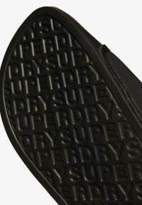 Zwarte rubberen sneakerzool met verhoogde tekst "SUPERDRY" in een patroonontwerp. Gladde randen en een slanke, gestroomlijnde vorm zijn zichtbaar.