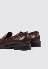 Zapatos de cuero marrón con punta redondeada, tacón bajo y costuras decorativas. Textura suave con acentos laterales y diseño de talón abierto.