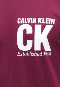 Bordowa bawełniana koszulka z białym napisem: "CALVIN KLEIN", "CK" oraz "Established 1968", o miękkiej fakturze i luźnym kroju.