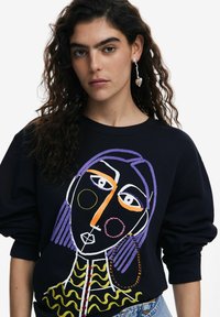 Sudadera negra con un diseño de cara bordada colorido y características abstractas. Patrón texturizado de volantes en la parte inferior. Mangas largas.