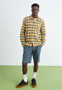 Chemise en flanelle à carreaux jaune, noir et blanc ; shorts gris ; baskets marron ; chaussettes blanches avec logo ; coupe décontractée, design à boutons sur le devant.