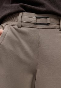 Zbliżenie na osobę w taupe'owych spodniach z wbudowanym materiałowym paskiem i metalową prostokątną klamrą, ręką schowaną w kieszeni, w zestawie z ciemnobrązową górą.