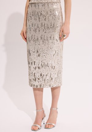 Srebrna połyskująca spódnica midi noszona przez osobę w srebrnych sandałach na wysokim obcasie oraz z dużym srebrnym pierścieniem na prawej ręce.