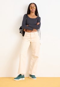 Haut noir à manches longues court, pantalon large crème, et baskets bleu sarcelle. Mannequin avec des cheveux tressés posant devant un fond uni.