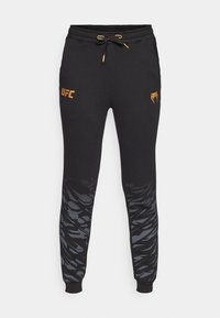 Sorte joggebukser med tapered fit, med elastisk midjebånd, snor, gyllen UFC-logo og et grått abstrakt mønster på de nedre ben.