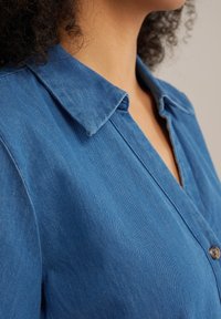 Zila denima krekla ar norādītu apkakli, pogu aizdari un gludu tekstūru. Audumam ir smalka auduma raksts.