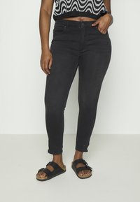 Jeans negros ajustados con un ligero estiramiento, con una cintura de talle medio, bolsillos frontales y dobladillos sin rematar, combinados con sandalias negras.