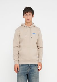Beige cotton hoodie with a front pocket, drawstring hood, and blue logo detail on the chest. Worn with blue denim jeans.