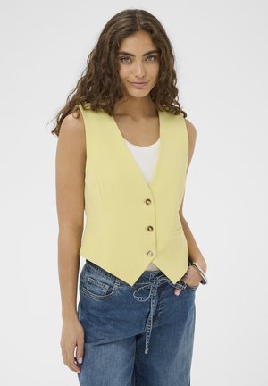 Frau mit lockigem Haar, die eine ärmellose gelbe Weste mit Knöpfen über einem weißen Oberteil und blauen Jeans trägt, steht mit einer Hand in der Tasche.