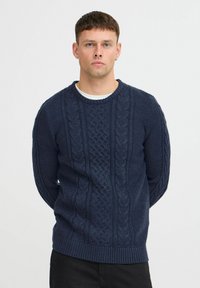 Pull en maille torsadée bleu marine avec des motifs texturés, col rond, poignets et ourlet côtelés. Présente divers points de tricot pour un détail supplémentaire.