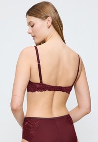 Nainen nähtynä selkäpuolelta, päällään saman sävyiset viininpunaiset pitsiliivit ja korkeavyötäröiset alushousut, vaaleaa taustaa vasten.