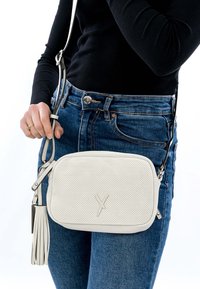 Hvid perforeret crossbody taske med kvast, båret over mørk langærmet top og blå jeans, hånd der justerer remmen.