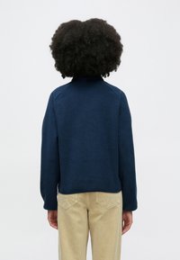 Marineblå fleece genserjakke med høy krage og raglanermer. Myk tekstur, kortest foran på kanten, kombinert med lyse khakibukser.