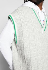 Graue Strickweste mit V-Ausschnitt, mit strukturierten Details und grünem Besatz entlang der Armöffnungen und des Ausschnitts. Über einem weißen Hoodie getragen.