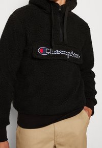Schwarzer Fleece-Pullover mit halbem Reißverschluss, ausgestattet mit einem auffälligen, gestickten "Champion"-Logo in Blau und Weiß auf der Brust sowie gerippten Bündchen.